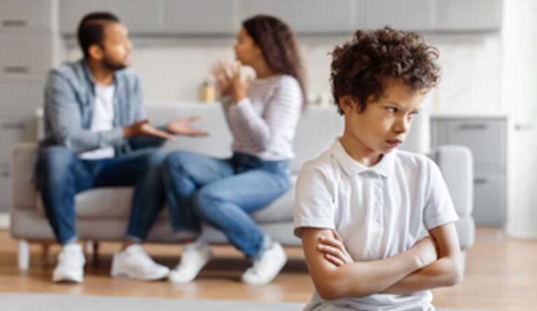 Menino triste com braços cruzados enquanto os pais discutem desfocados ao fundo em ambiente doméstico, representando impacto emocional do conflito parental | Valente Perícias